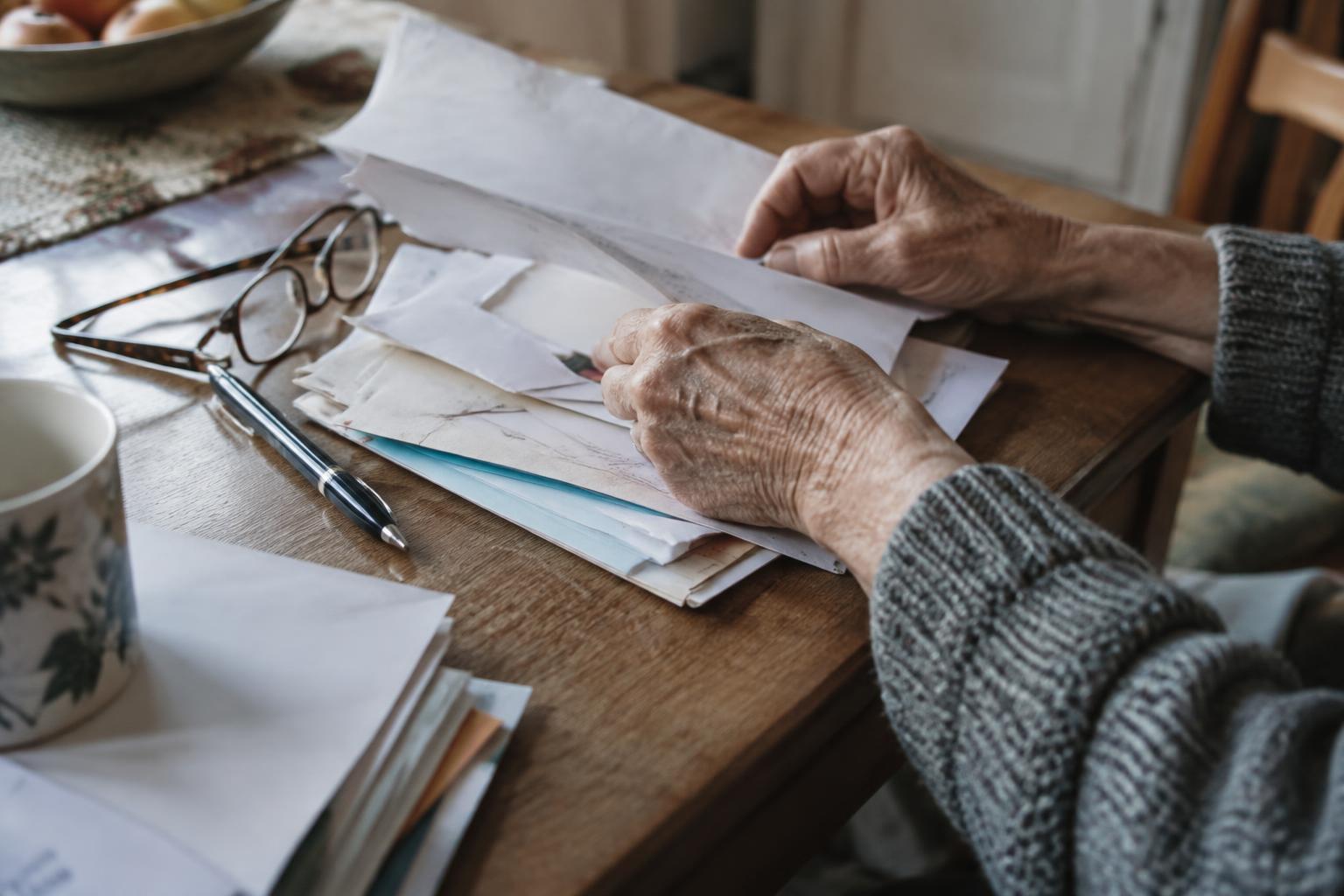 Close-up of an older person's hands sorting through a stack of letters and paperwork at a kitchen table, with reading glasses and a pen beside them
